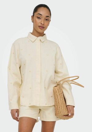 Femme portant une chemise crème boutonnée et un short assorti avec de petits motifs de cœur, tenant un sac à main en paille tressée.