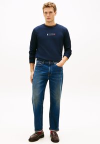 Camicia a maniche lunghe blu navy con logo, abbinata a jeans blu a gamba dritta e scarpe marroni. Presenta polsini arrotolati e calzini di colore contrastante.