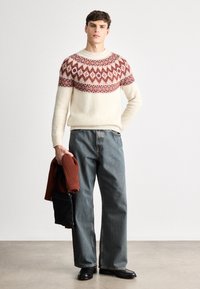 Strickpullover in Creme mit rotem geometrischen Muster an der Passe. Lockere Jeans aus Denim, hält eine rostfarbene gerippte Strickjacke und eine schwarze Tasche.