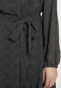 Svart klänning med vita prickar, med ett knytbälte i midjan, långa ärmar med elastiska manschetter och en lätt, genomskinlig textur.