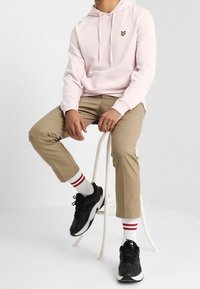 Hellrosa Kapuzenpullover mit kleinem gelbem Logo, beige Hose, schwarze Turnschuhe und weiße Socken mit roten Streifen, sitzend auf einem weißen Hocker.