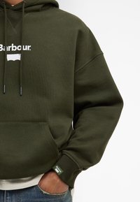 Homme portant un sweat à capuche Barbour vert olive avec poche avant et une petite étiquette Barbour sur la manche, main dans la poche, chemise blanche en dessous.