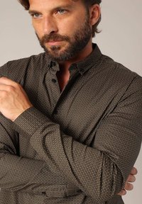 Bruine overhem shirt met een subtiel wit stippenpatroon, lange mouwen, knoopkraag en textuurstof. Aangedreven met zwarte knopen.