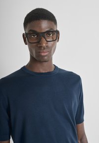 Maglione lavorato a maglia blu navy, a maniche corte e con collo a giro; il modello indossa occhiali rettangolari neri, mostrando una texture liscia e un design aderente.