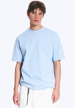 Tânăr cu păr castaniu scurt, purtând un tricou bleu cu mânecă scurtă și pantaloni albi pe un fundal alb simplu.