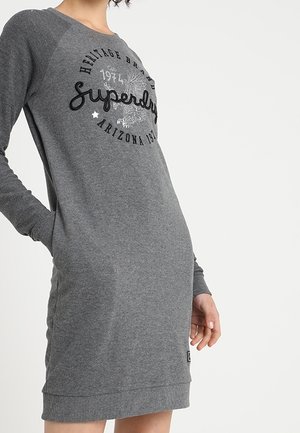 Kobieta ubrana w szarą sukienkę-swetrową z długim rękawem, z napisem "Superdry 1974 Arizona" i grafiką orła na przodzie.