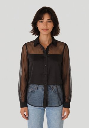 Donna con capelli corti e scuri che indossa una camicetta nera trasparente a maniche lunghe con bottoni e jeans azzurri, in piedi davanti a uno sfondo neutro.