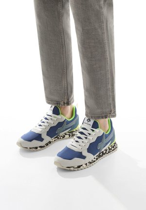 Blaue und graue Sportschuhe mit weißen Schnürsenkeln, einer strukturierten Sohle mit einem schwarz-weißen Muster und grünen Akzenten an der Ferse und am Kragen.