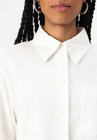 Camicia bianca con bottoni realizzata in cotone; presenta un colletto classico, tasca frontale e quattro chiusure a bottone. Texture liscia, design minimalista.