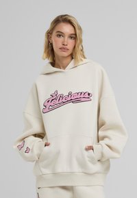 Felpa oversize color crema con logo ricamato rosa "Felicieux", tasca a marsupio e dettagli a forma di farfalla sulle maniche. Tessuto morbido, vestibilità comoda.
