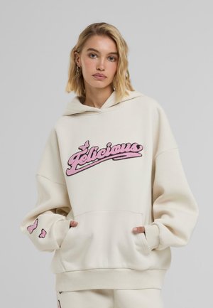Sweat à capuche crème surdimensionné avec logo "Felicieux" brodé en rose, poche kangourou et accents de papillon sur les manches. Tissu doux, coupe décontractée.