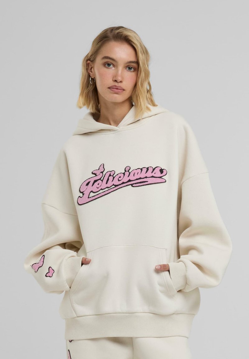 Sweat à capuche crème surdimensionné avec logo "Felicieux" brodé en rose, poche kangourou et accents de papillon sur les manches. Tissu doux, coupe décontractée.