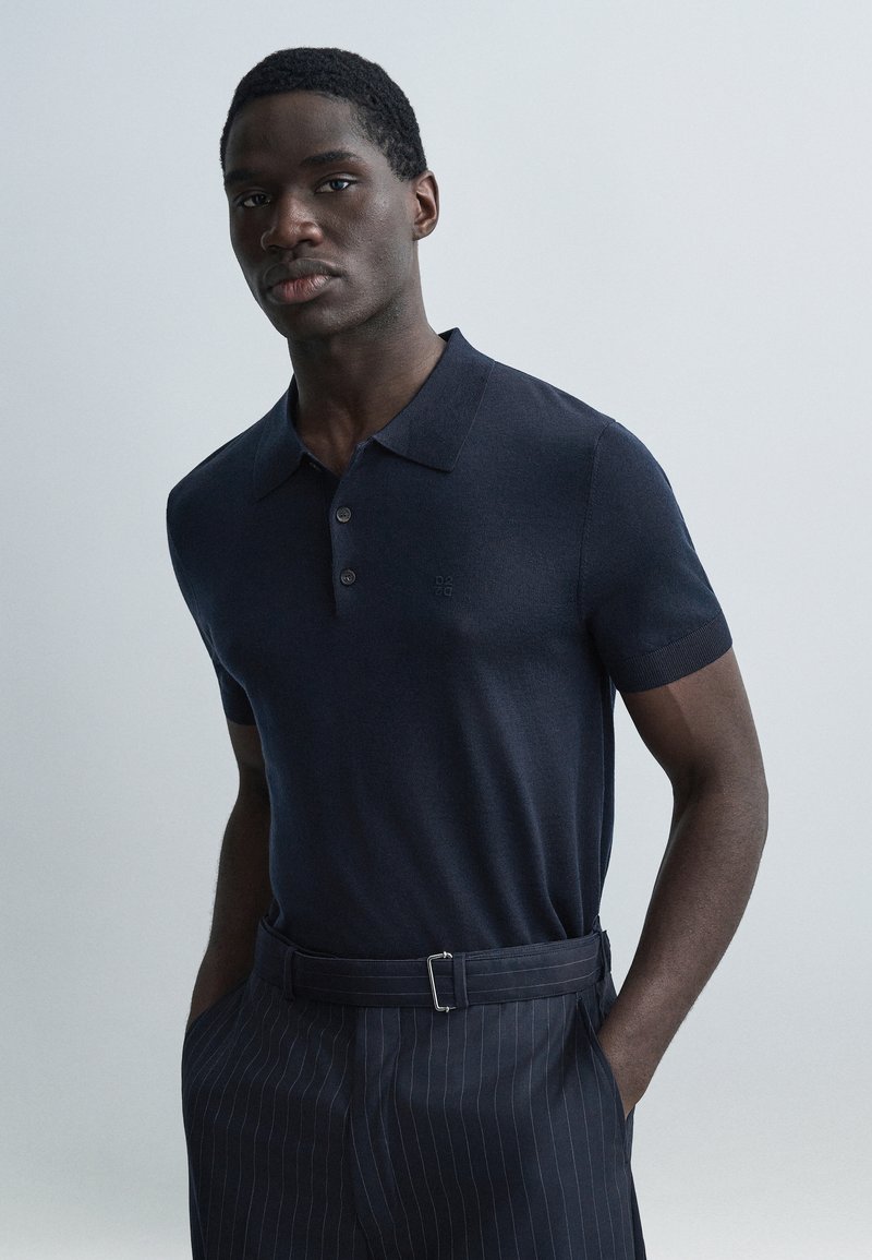 Μπλε ναυτικό πόλο μπλουζάκι με κοντά μανίκια, κατασκευασμένο από υφασμάτινη υφή, με πατιλέτα τριών κουμπιών και διακριτικό λογότυπο. Φοράται με παντελόνια με λεπτές ρίγες.