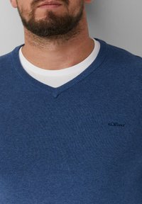 Dunkelblauer V-Ausschnitt-Pullover aus Strickstoff, ausgestattet mit einem kleinen Logo auf der linken Seite und über einem weißen Rundhalsausschnitt-T-Shirt getragen.