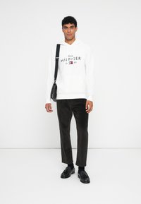 Hvit hettegenser med "Tommy Hilfiger"-trykklogo, svarte cordbukser og svarte loafers. Modellen holder en liten svart skulderveske.