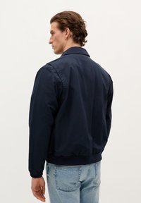 Marineblå, letvægts bomberjakke med krave, elastiske manchetter og talje. Båret over lyseblå jeans, set fra ryggen.