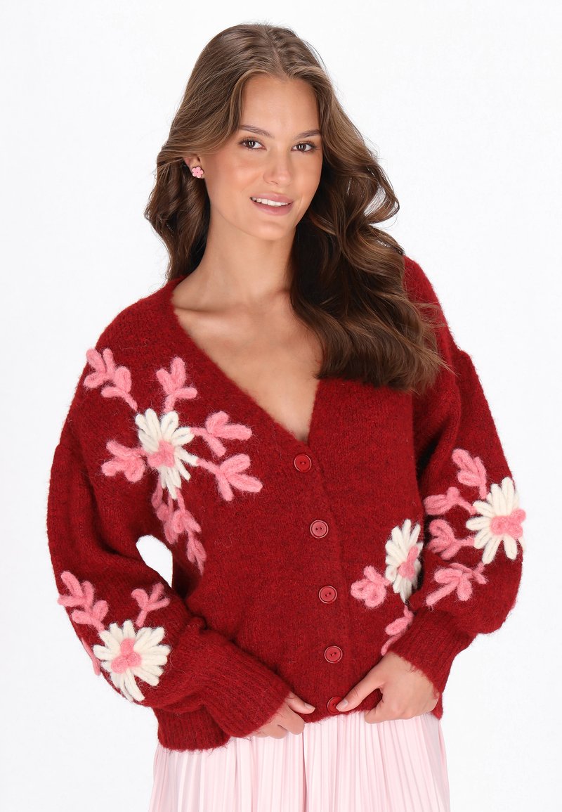 Röd ullkofta med djup V-ringning, dekorerad med rosa och vita blommiga broderier på ärmarna, samt fem runda knappar nedtill fram.