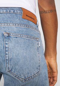 Close-up de uma pessoa a usar jeans de ganga azul claro com um rótulo de couro Woodbird e um antebraço tatuado visível.