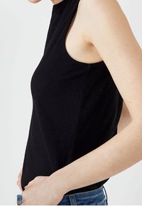 En slank person iklädd en ärmlös svart topp och blå jeans, som står sidledes med händerna vilande på höfterna mot en enfärgad vit bakgrund.