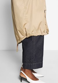 Alara jalat pukeutuneita tummansinisiin croppattuihin farkkuihin, beigessä takissa jossa on narureuna, ja valkoisissa avosandaaleissa keskikorkeat korot harmaalla lattialla.