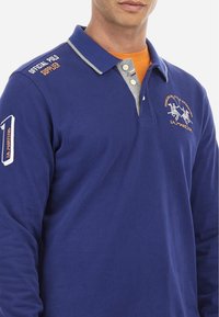 Långärmad blå pikétröja med grå krage, med broderad logotyp och text på bröstet och ärmen. Knappar i en kontrasterande färg.