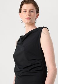 Black sleeveless dress featuring a draped neckline and soft texture, with ruching on the side. Model wears silver hoop earrings.