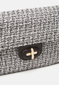 Pochette en tissu tweed noir et blanc avec une bordure en cuir noir et une fermeture à tourniquet rectangulaire dorée sur un fond clair.