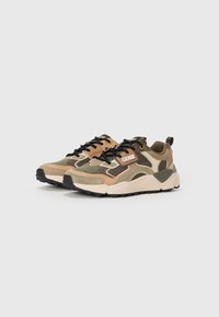 Las zapatillas de camuflaje presentan una mezcla de materiales en beige, verde y marrón, con una suela gruesa y cordones negros. Los acentos notables incluyen un parche con el logo.