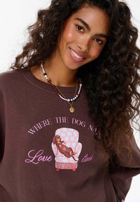 Hnědá mikina s růžovým grafickým textem a ilustrací spícího psa na pastelově barevné židli, se květinovými vzory a červenými akcenty.