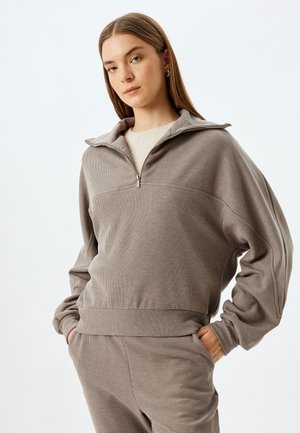 Szary, strukturalny sweatshirt z wysokim kołnierzem i zamkiem błyskawicznym. Luźny krój z ściągaczami przy mankietach i u dołu. Noszony z pasującymi szarymi spodniami.