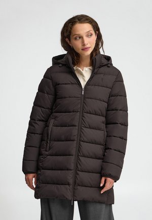 Donna che indossa un cappotto invernale imbottito nero con cappuccio e cerniera frontale, in piedi davanti a uno sfondo chiaro e semplice.