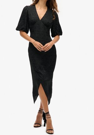 Woman wearing a black midi dress with puff sleeves, V-neck, asymmetrical slit, and black strappy high-heeled sandals, standing with hands clasped.
