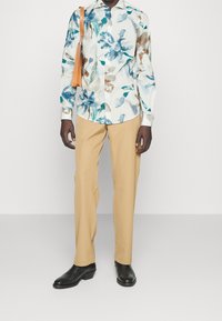 Chemise à manches longues à motif floral blanche avec des accents bleus et bruns, associée à un pantalon beige et des chaussures noires. Une main tient un sac beige.