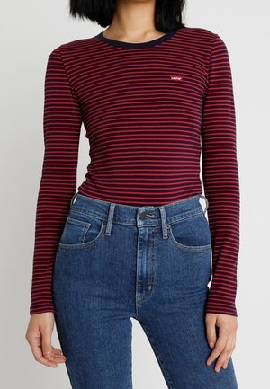 Mujer con una camiseta de manga larga a rayas rojas y azul marino metida en unos vaqueros de talle alto azules, de pie contra un fondo liso.