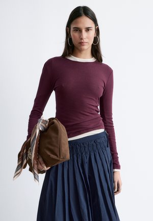 Kvinde med langt, mørkt hår iført bordeaux langærmet top, navy plisseret nederdel, store hoop-øreringe og bærende på en brun taske med mønstret tørklæde.