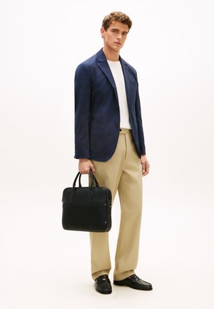 Giovane uomo con blazer blu navy, camicia bianca, pantaloni beige, mocassini neri, che tiene una borsa da lavoro in pelle nera, in piedi in uno spazio neutro.