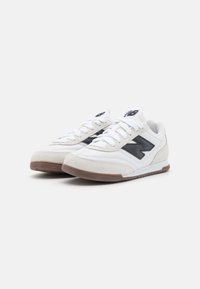 Pari valkoisia ja beigejä New Balance -lenkkareita mustalla logolla ja ruskeilla pohjilla, vinosta sivukuvasta valkoista taustaa vasten.