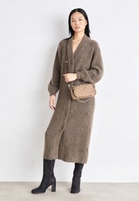 Donna che indossa un lungo cardigan di maglia marrone, stivali neri con tacco e una borsa a tracolla beige con un design a motivo, in piedi contro un muro bianco semplice.