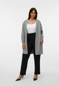 Lichtgrijze cardigan met lange mouwen en zijzakken, gedragen over een witte top, gecombineerd met zwarte broeken en puntige zwarte hakschoenen.