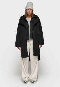 Parka nera impermeabile con un look a strati, abbinata a una giacca imbottita grigia, pantaloni a gamba larga crema e un berretto grigio. Sneakers argento.