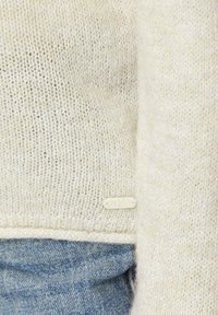 Lichtbeige gebreide trui met een gestructureerd patroon, een geribde zoom en een klein wit label met "QS" nabij de onderkant. G gedragen over lichtblauwe denim.