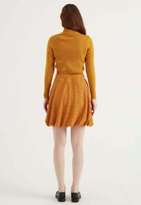 Turtleneck a coste giallo senape abbinato a una mini gonna a zigzag con texture in una tonalità simile. Scarpe nere slip-on completano il look.