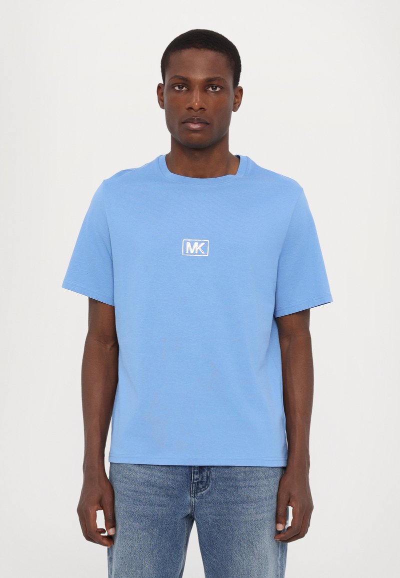 Lyseblå bomull T-skjorte med korte ermer, rund hals og hvit MK-logo på brystet. Båret med mellomvasket denimbuks.