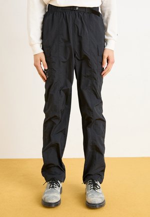 Pantalons cargo noirs avec une taille élastique et une ceinture. Dispose de plusieurs poches latérales et d'un tissu léger et texturé. Portés avec des chaussures grises.