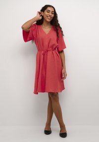 Femme aux longs cheveux bouclés portant une robe corail à mi-mollet nouée à la taille, posant avec une main près de l'oreille et des ballerines noires.
