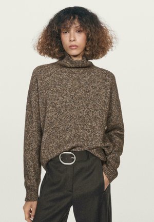 Pull en maille marron à col roulé avec une coupe décontractée, motif texturé. Associé à un pantalon noir sur mesure et une grande boucle de ceinture en argent.