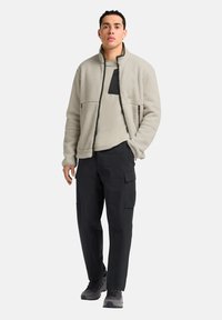 Fleece jas in lichtgrijs met een hoge kraag, rits aan de voorkant en twee ritszakken; gecombineerd met een beige shirt en zwarte cargo broeken.