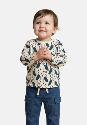 Tout-petit aux cheveux bruns souriant, portant un t-shirt à manches longues avec un imprimé macareux bleu marine et rouge et un pantalon cargo bleu, les mains jointes.