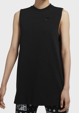 Persoon draagt een mouwloos zwart Nike-shirt met ronde hals en zwart-witte legging met patroon, staand tegen een effen achtergrond.