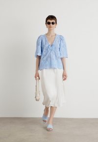 Lichtblauw kanten top met puffmouwen, witte satijnen rok, lichtblauwe instappers en een crèmekleurige tas. Neutrale achtergrond.